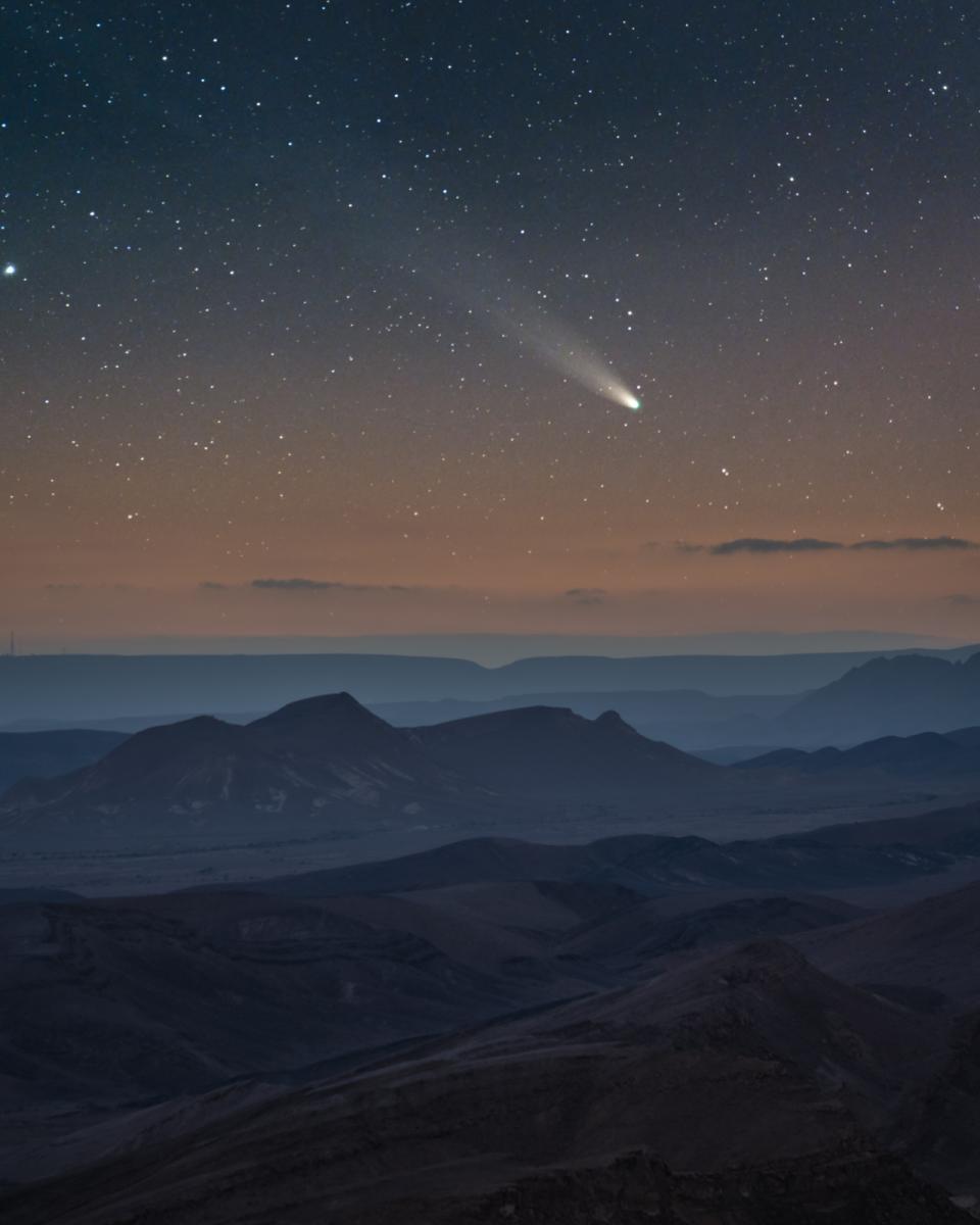 Photograph of a comet entering the Earth's atmosphere. A rocky landscape occupies the bottom half of the frame, while the bright white tail of the comet can be seen moving across the sky above