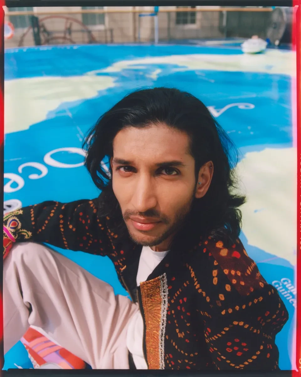 Portrait photograph of a young man with brown skin, a closely trimmed black beard and long black hair. He is sitting on the ground, his face turned upwards towards the camera