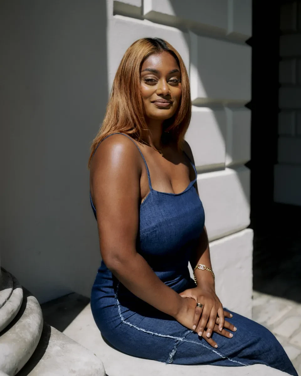 Portrait photograph of a woman with dark brown skin and long straight hair. She is sitting on a low wall, her face smiling and turned towards the camera