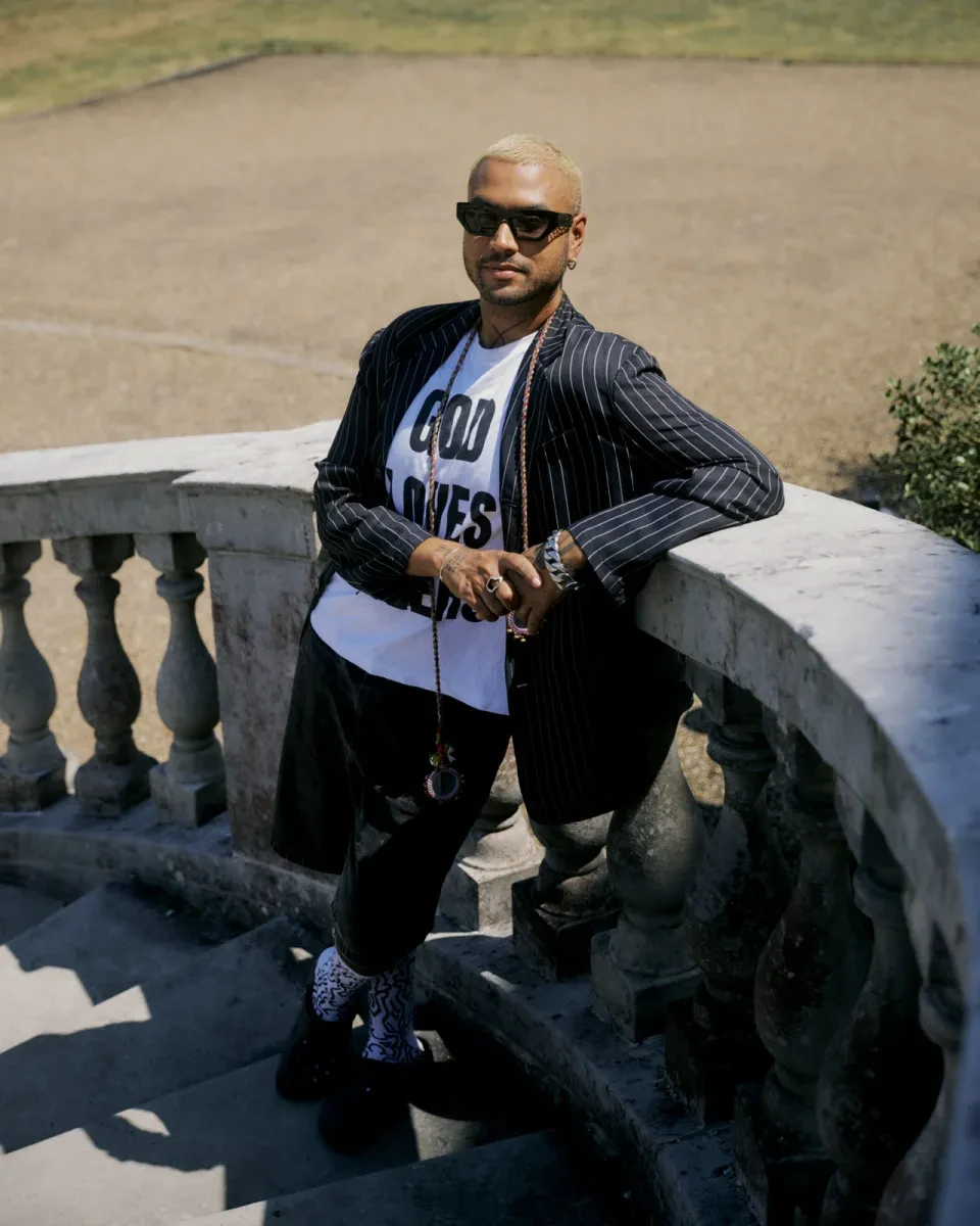 Portrait photograph of a man standing outside on a grand stone staircase. He wears a pin-striped suit jacket, white t-shirt with a half-concealed slogan on it and thick black sunglasses