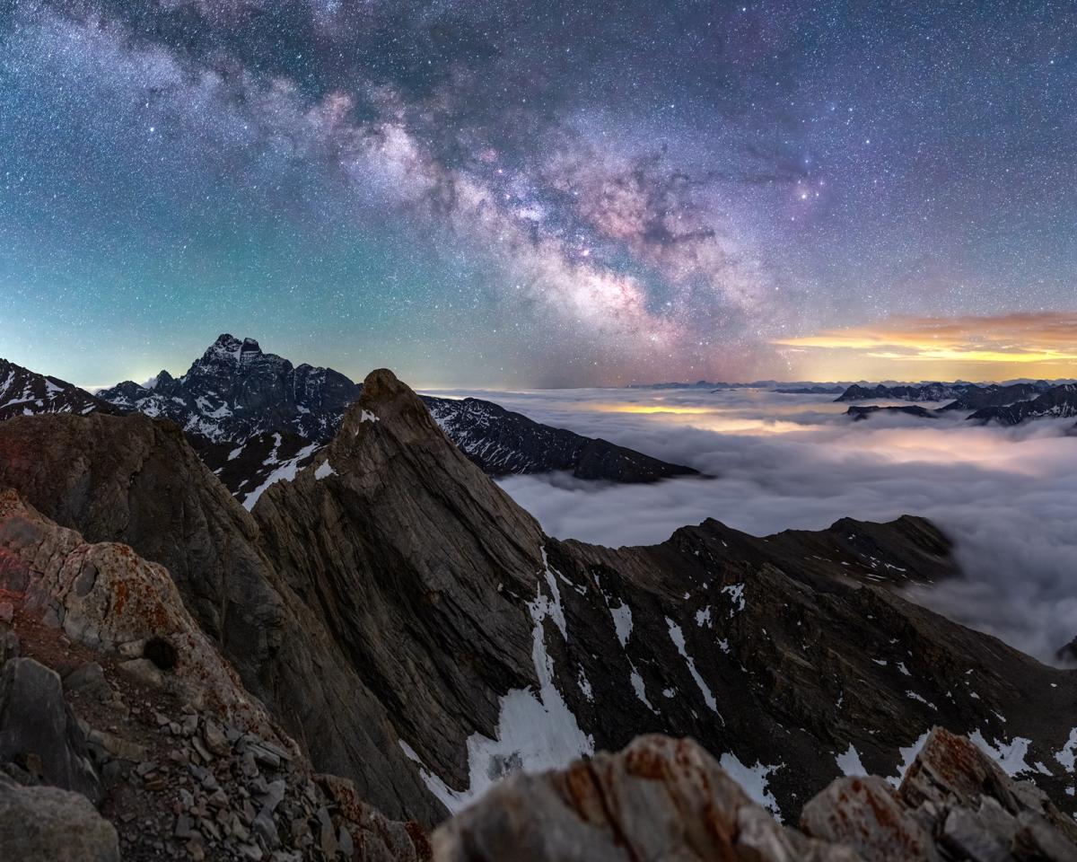 View of rocky top of a mountain with the Milky Way diagonal in the sky behind, with the sky in tones of purples, blues and yellow, with fog at the top of the mountain