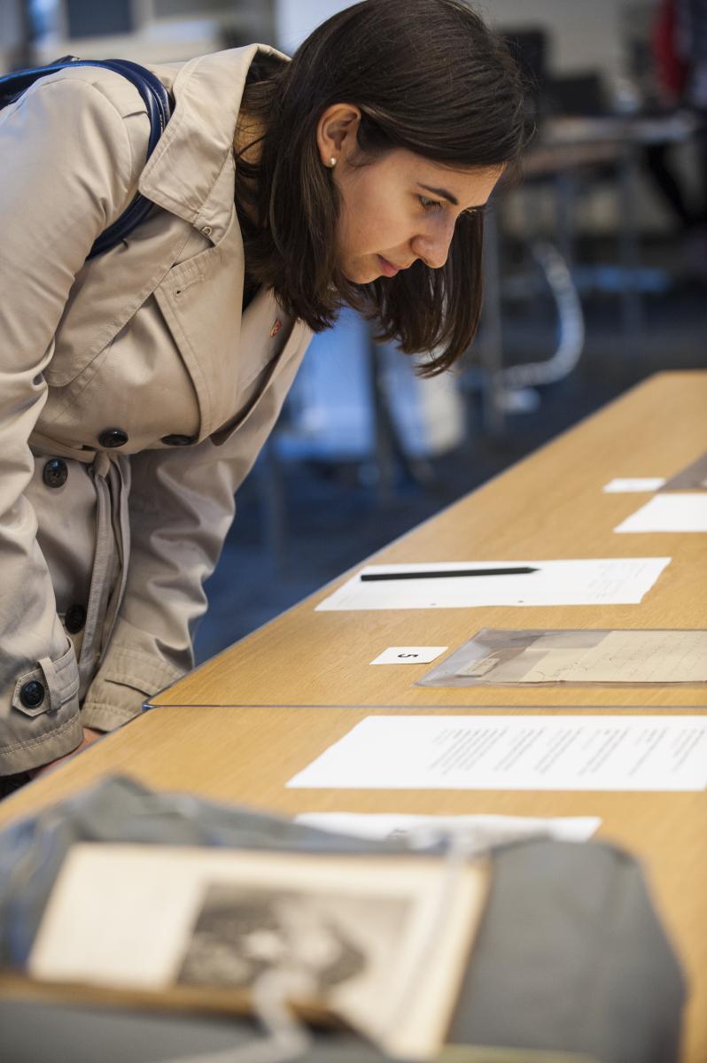 A woman in a cream trench coat looks at a display of manuscripts and books on a table