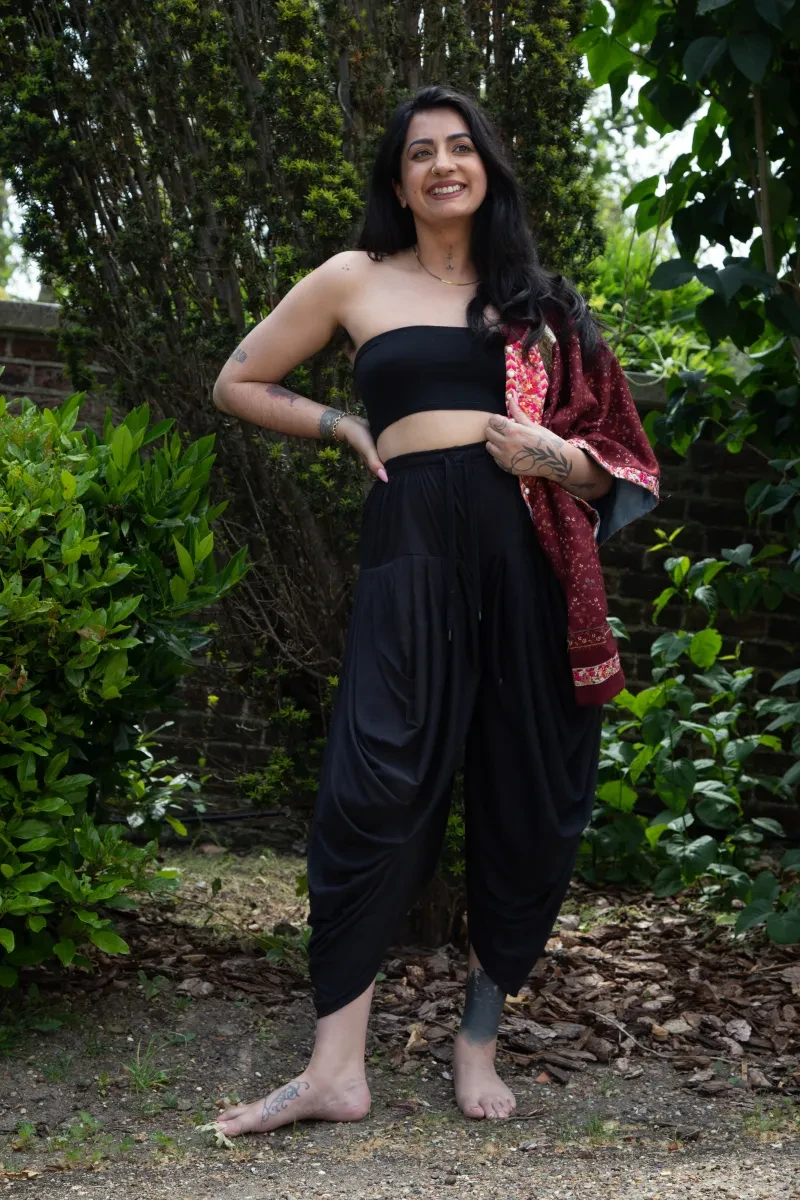 A photo of Sharan Dhaliwal who stands in front of vibrant greenery, wearing a black cropped top and high waisted black cropped trousers and a pink dupatta over one shoulder
