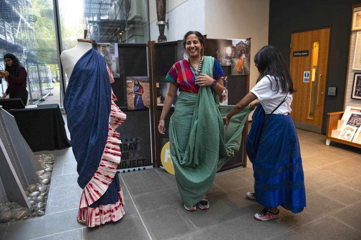 Image of woman helping another woman put on a green sari, the woman wearing the sari is smiling and happy while the other handles the fabric