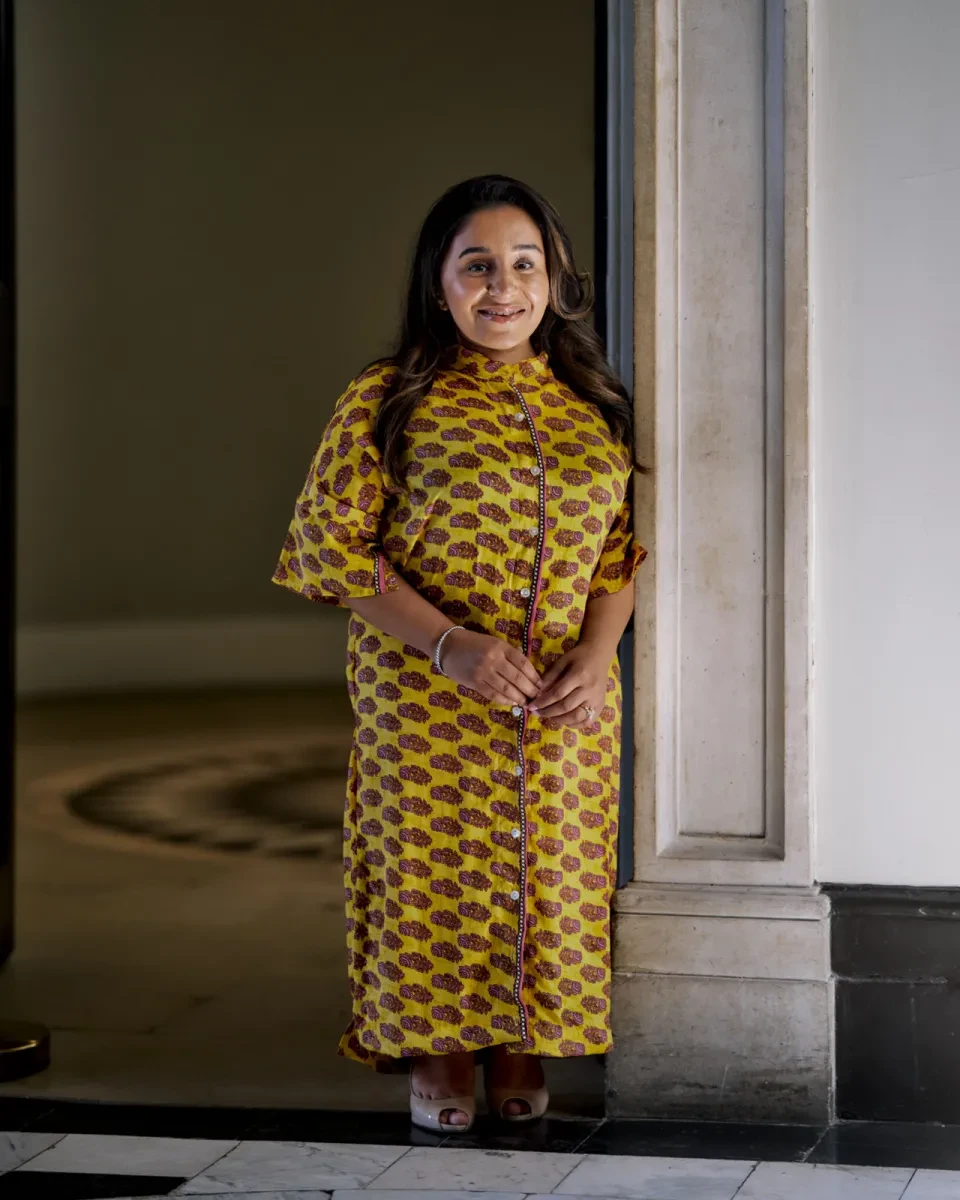 Portrait photograph of a woman wearing a brightly patterned yellow dress. She is standing in the doorway of an historic house and smiling straight at the camera