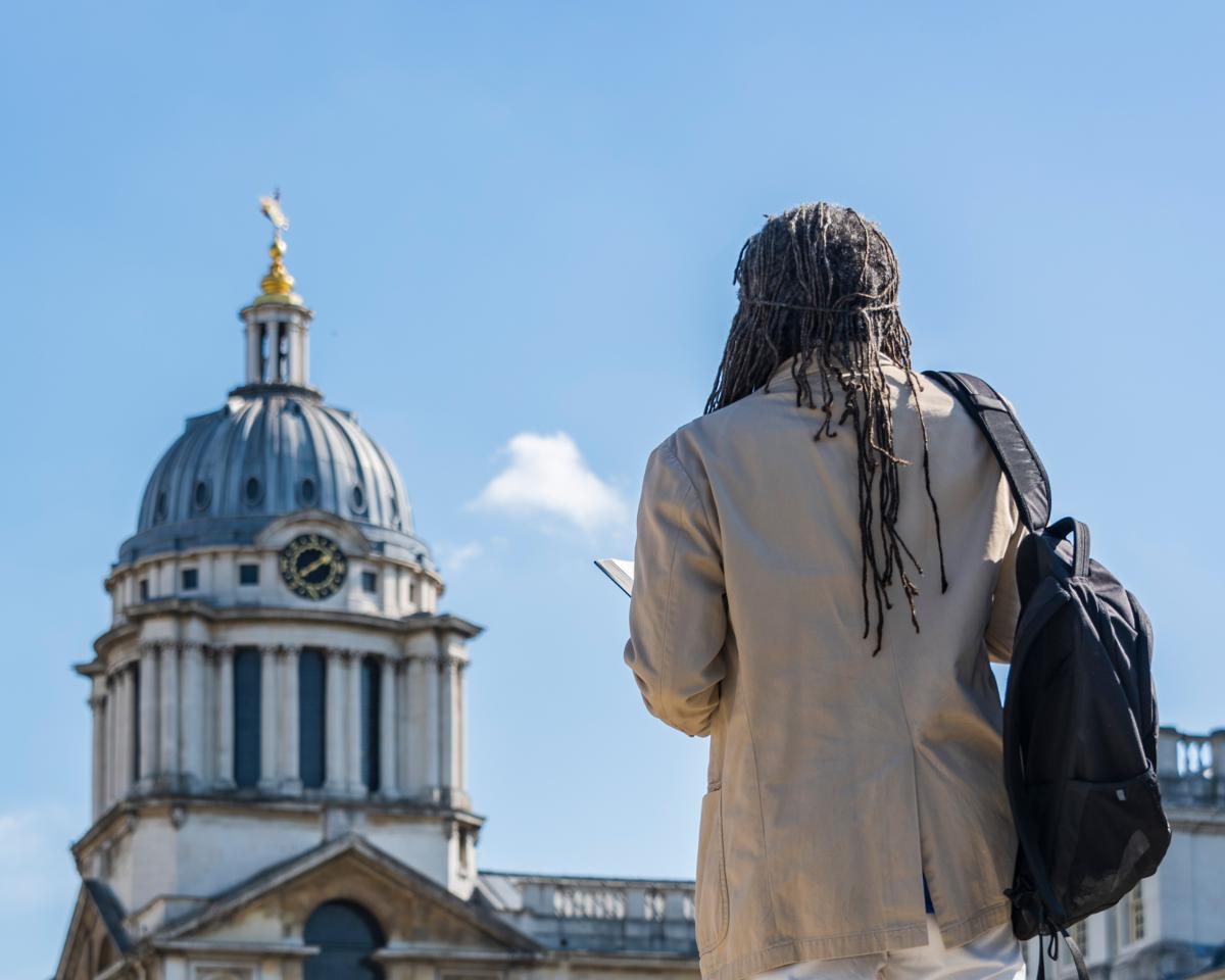 A man stands with his back to camera, looking up at a large round clock tower, part of the Old Royal Naval College building in Greenwich