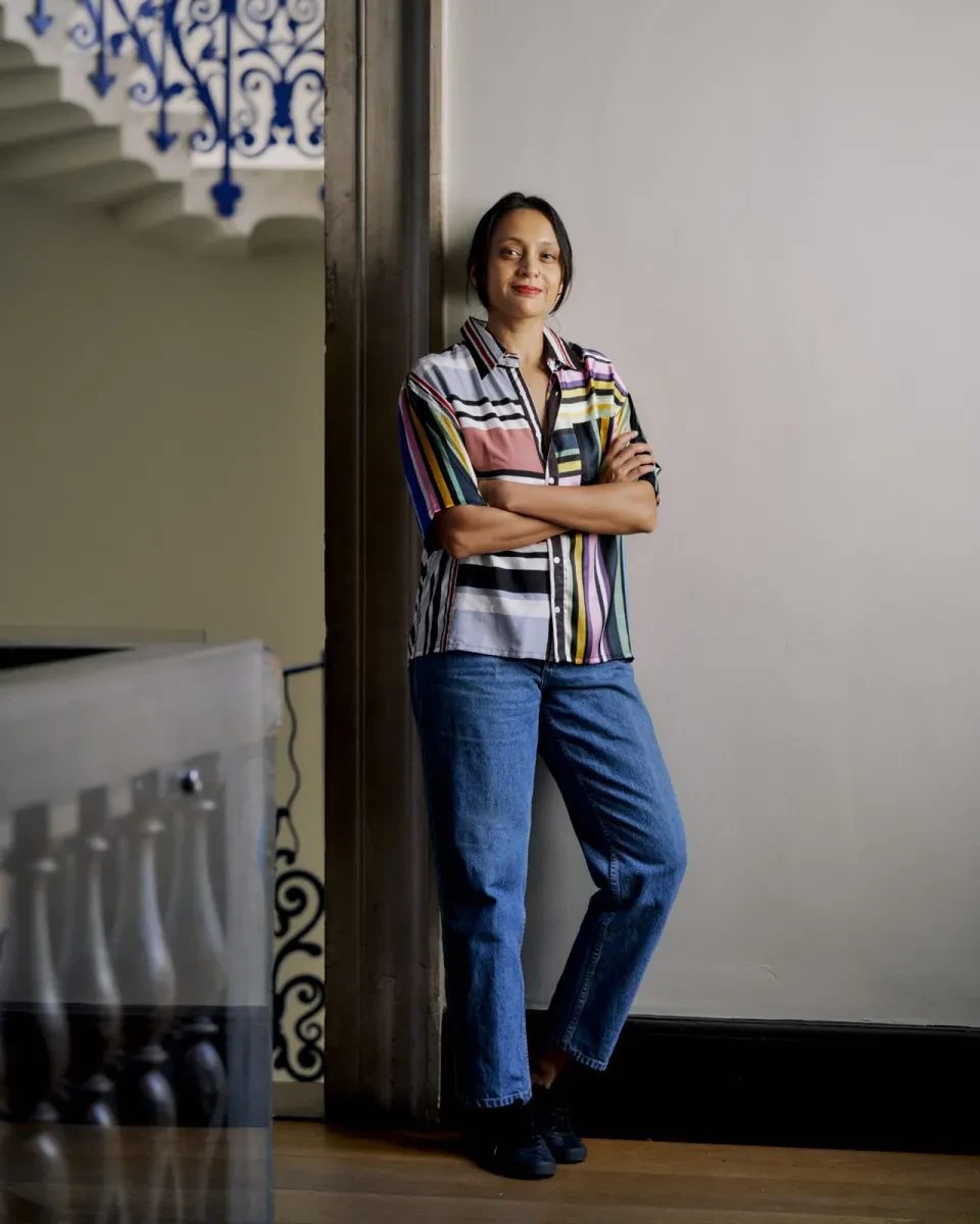 Portrait photograph of a woman leaning against a wall. She has her arms folded, and is smiling confidently at the camera.