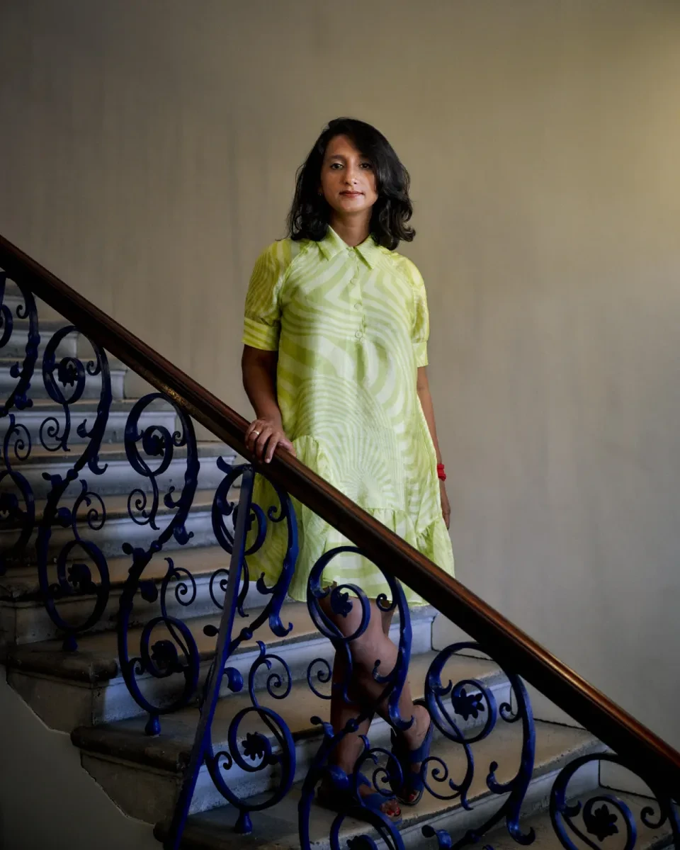 Portrait photograph of a woman wearing a pale yellow patterned dress. She is standing part-way down an ornate staircase, resting her hand against the bannister and looking directly at the camera