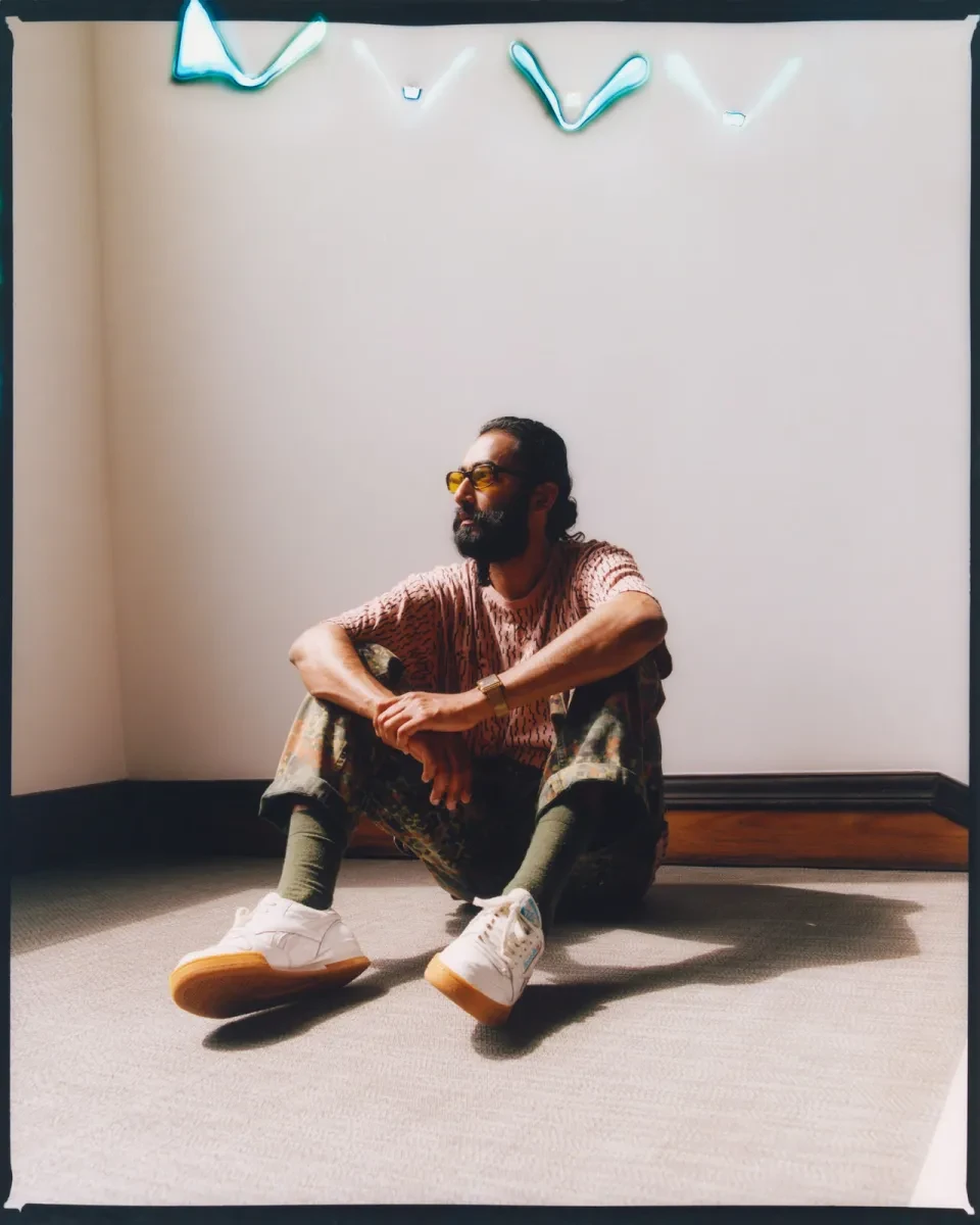 Portrait photograph of a man sitting on the ground in part-shadow. He has a thick black beard, wearing yellow-tinted glasses and a pale pink patterned t-shirt. He's gazing off to the right, arms resting on his knees 