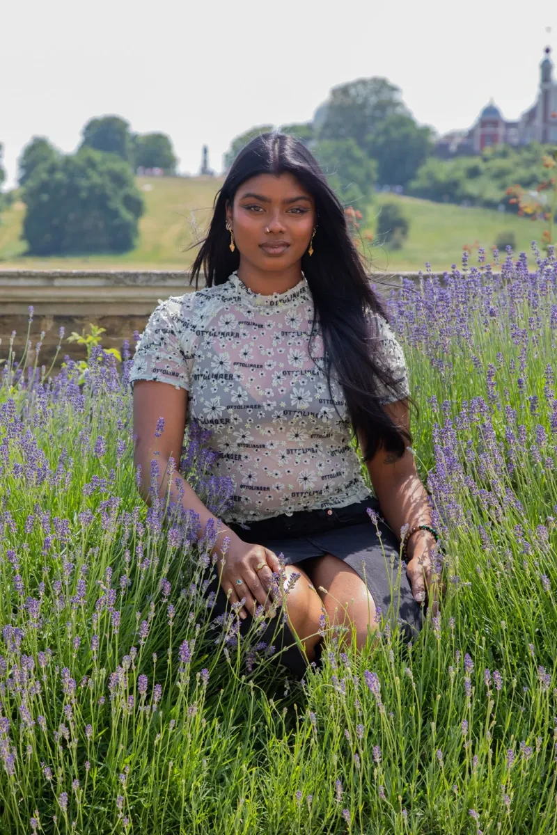 A woman kneels amidst lavender bushes, with Greenwich Park in the background. She has dark brown skin, long straight black hair and a neat nose ring and gold earrings. She is looking straight at the camera