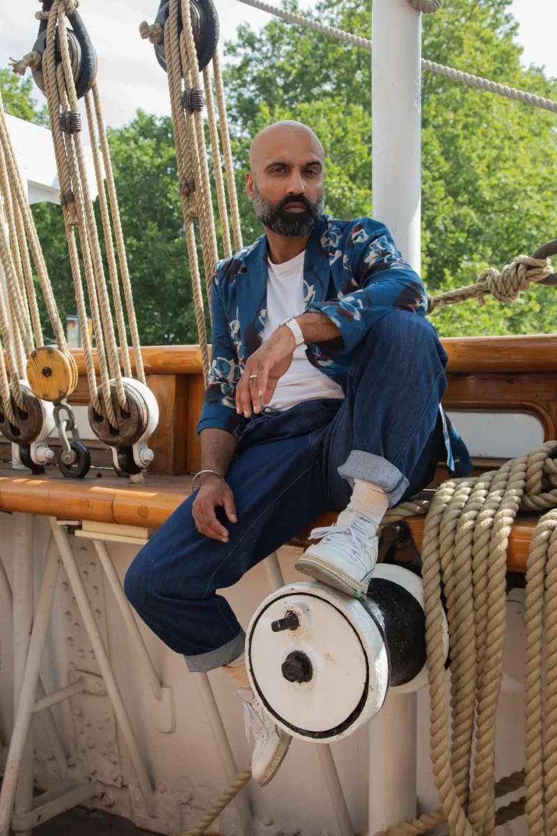 A photo of Taran Wilkhu sitting casually on the side of the Cutty Sark with lines of ropes and the tops of trees in the background. Taran wears denim trousers, a denim short with a white-shirt underneath and white trainers. Taran has a full moustache and beard. 