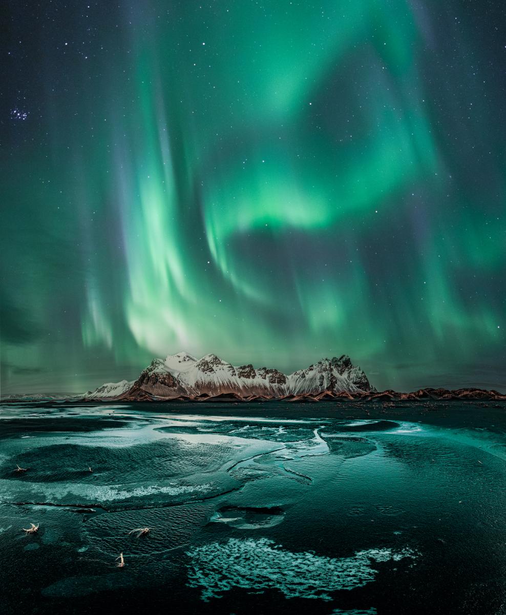 Image of the Vestrahorn in Iceland with the aurorae in the sky above, in teals and turquoise tones. The aurorae are reflected in the water that fills the bottom of the image.