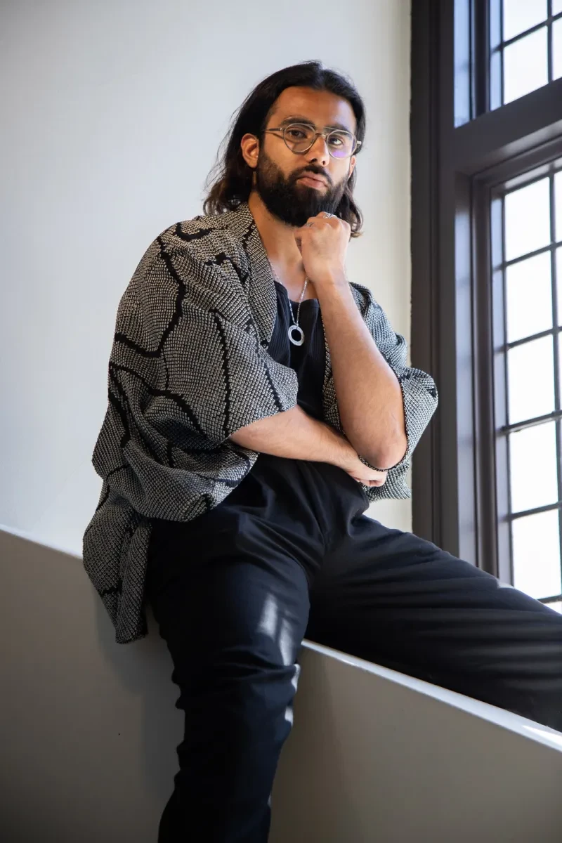 Portrait photograph of a man straddling a low wall. He has brown skin, a trimmed black beard and long dark hair