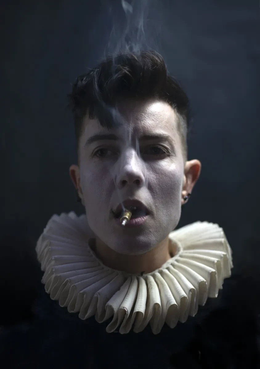 A portrait photo of E.M. Parry wearing a white ruff around their neck, their face painted white smoking a clay pipe with the smoke rising in a line in front of their face