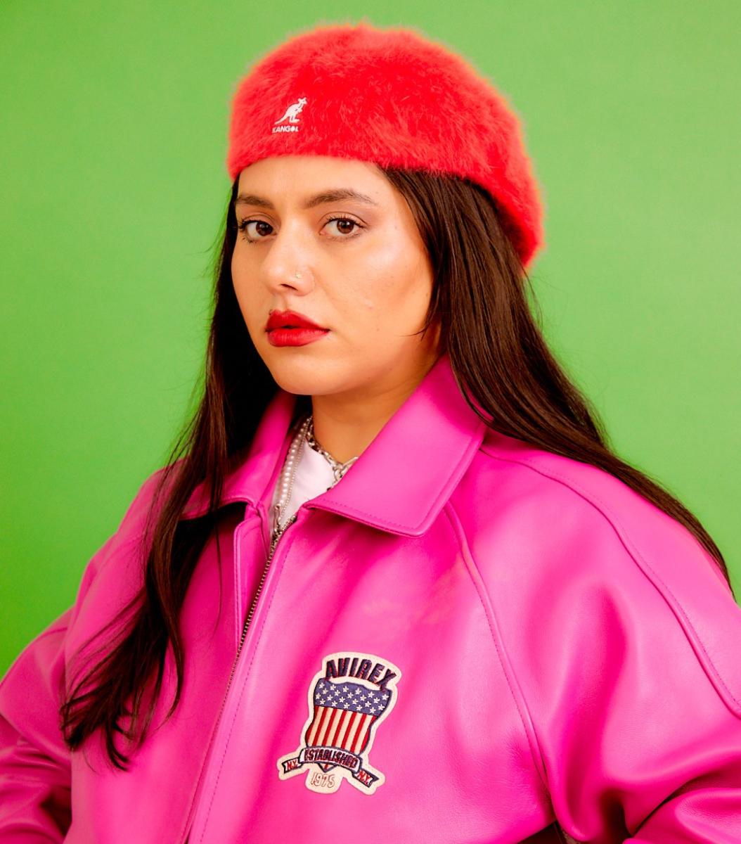 Image of Tara Kumar wearing a fluffy red hat and a pink bomber jacket against a green background
