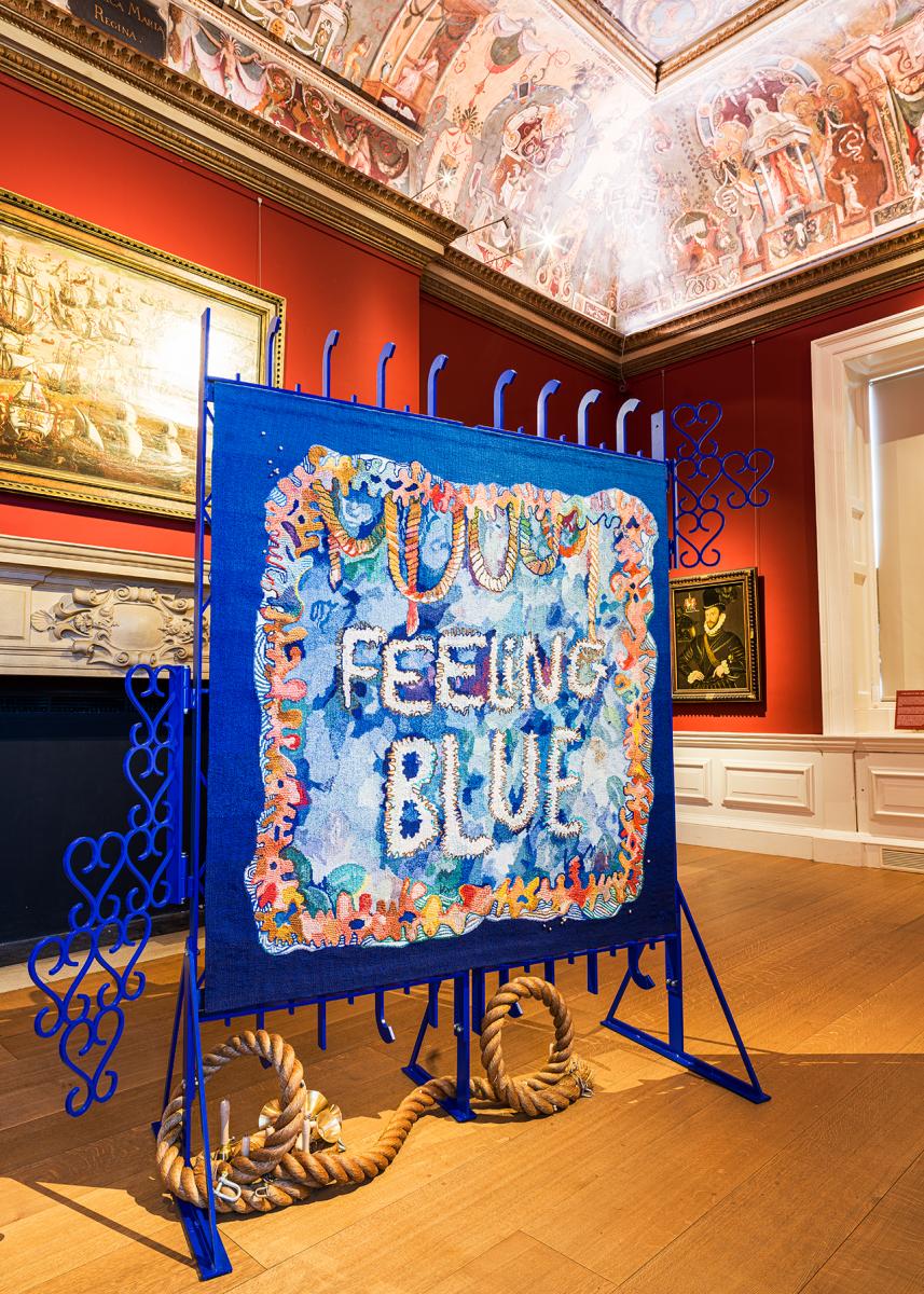 The artwork Feeling Blue on display in the Queen's House. It is a tapestry hung on blue metal gates, with a bright design in the centre of which are the words 'Feeling Blue'