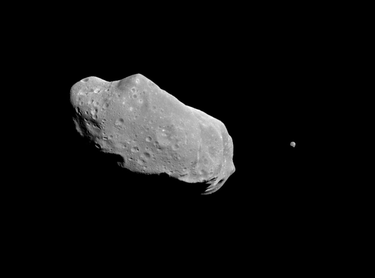 Image of a slightly oval shaped rocky asteroid in black space, with a small grey dot to the right which is its moon Dactyl