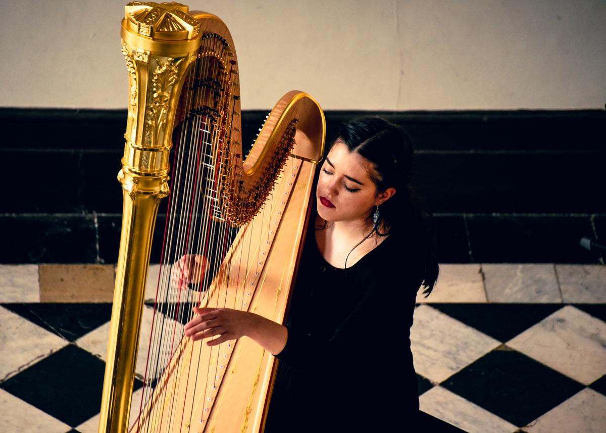 Photograph take from above of a woman playing a golden harp. She is wearing a black dress and an ornate black and white marble floor is in the background