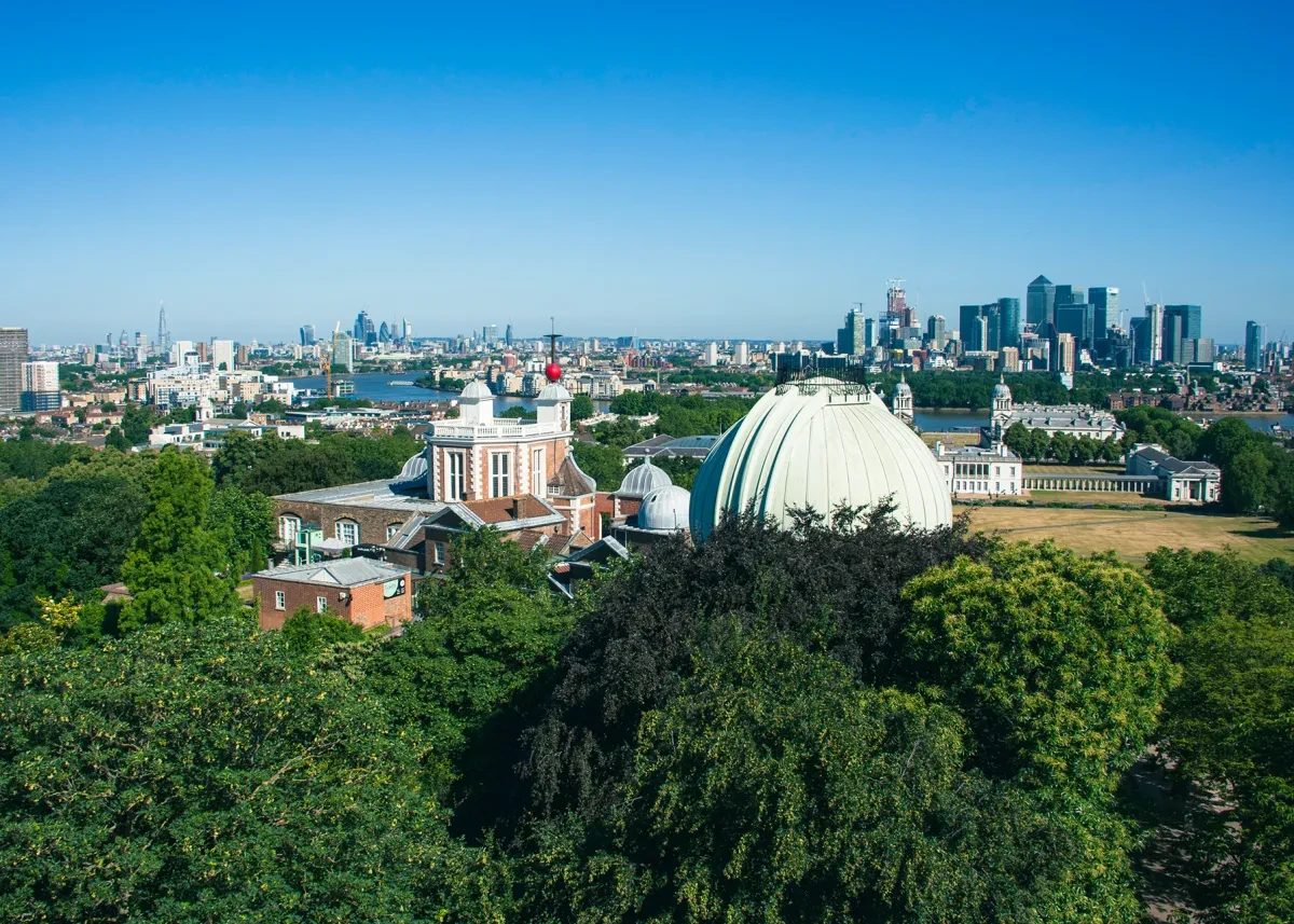 An aerial view of the Royal Observatory Greenwich, looking across the observatory domes and historic buildings towards the River Thames, with the London skyline in the distance