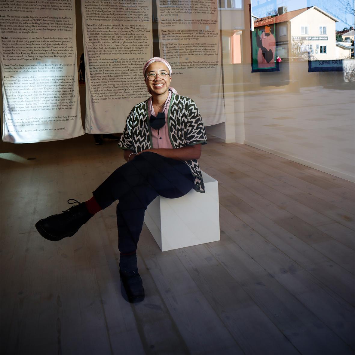 A photo of artist, Rudy Loewe, sitting in the middle of a gallery space on a white block. Rudy has joyful a smile and wears black trousers, a black and white short-sleeved jacket, a pink shirt and matching pink bandana.