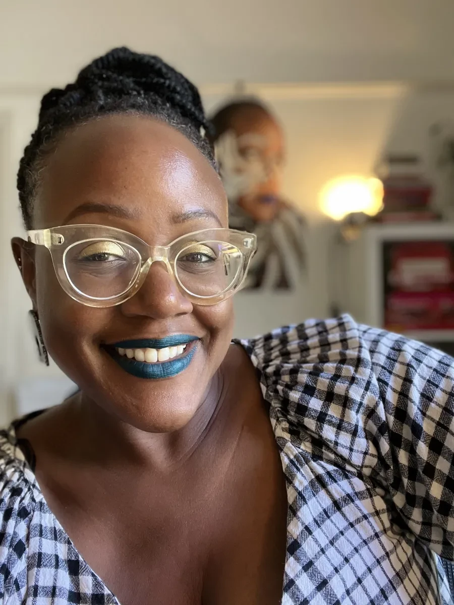 A photo of artist, Sharon Walters, who is Black woman smiling with blue lipstick and wearing translucent framed glasses and a gingham dress. Her hair is braided and tied up.