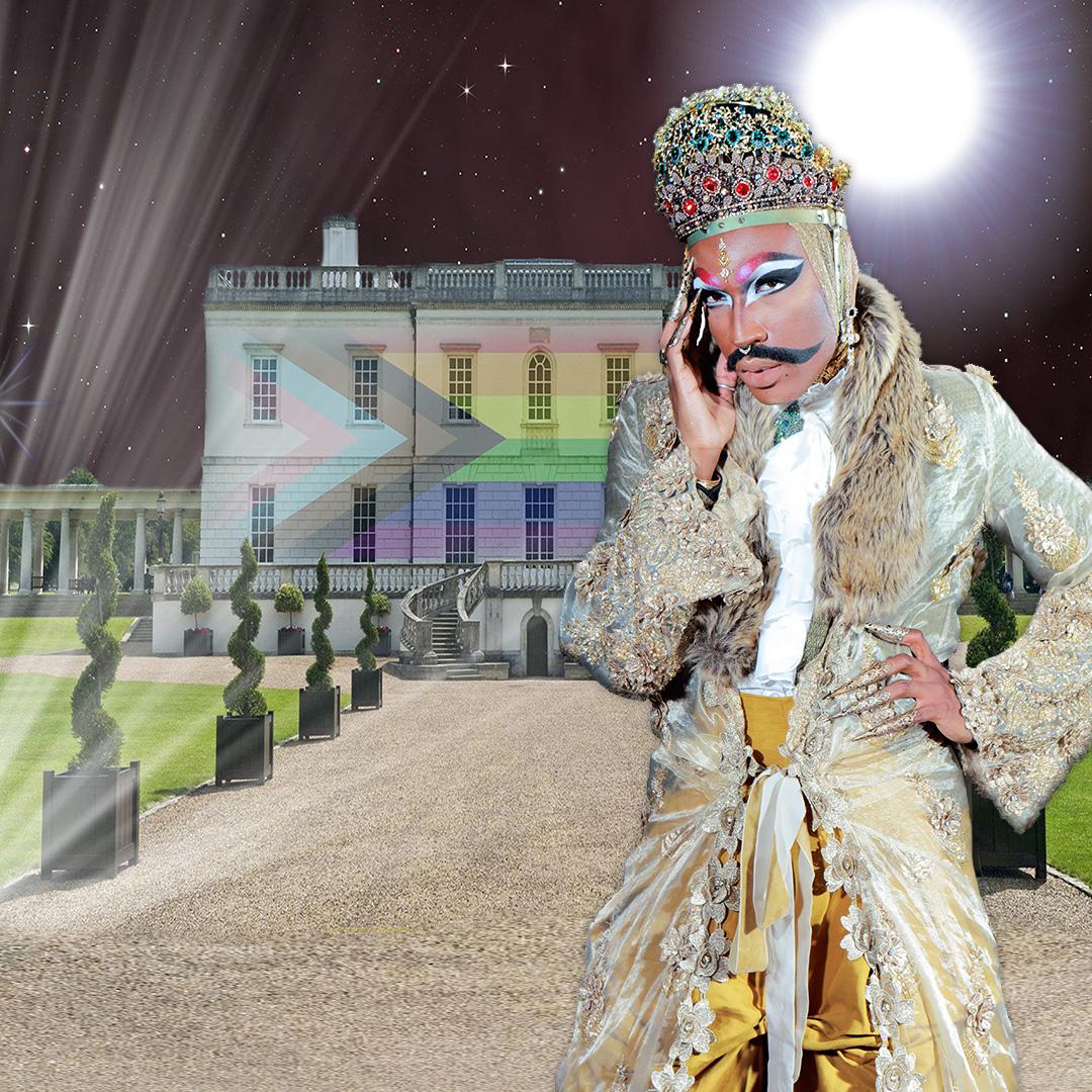 Rudy Jeevanjee stands in front of the Queen's House that's illuminated with the Progress Pride flag. He wears an opulent fur-trimmed and embroidered robe with a large jewelled crown. 