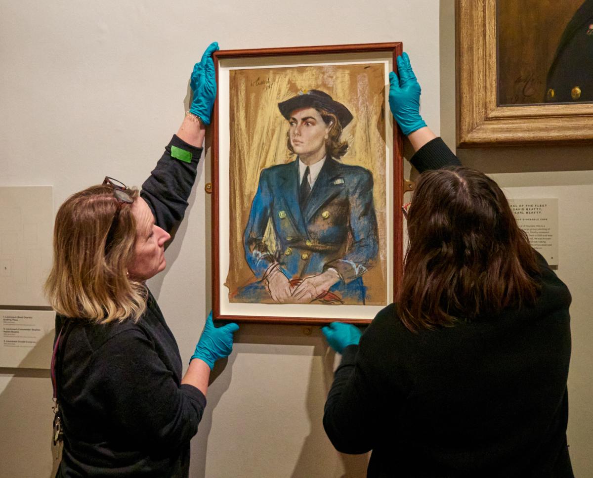 Two art object handlers hold a painting against a wall