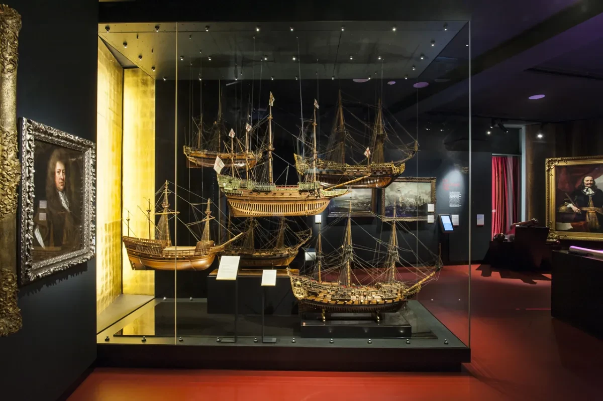 Image of 6 tudor and stuart ship models hanging inside a large glass case, on the side and behind you can see a number of paintings and portraits on the walls