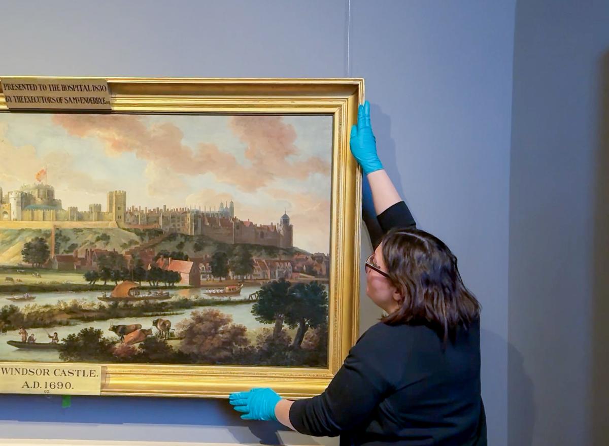 A woman holds a frame for display in an art gallery. She is wearing blue gloves to protect the painting's frame