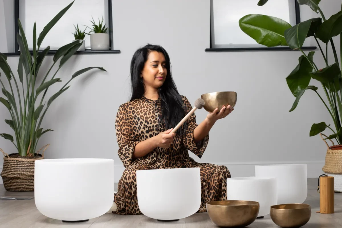 a photo of a woman sitting on the floor, with a bowl in hand and using an instrument to create a sound bath 