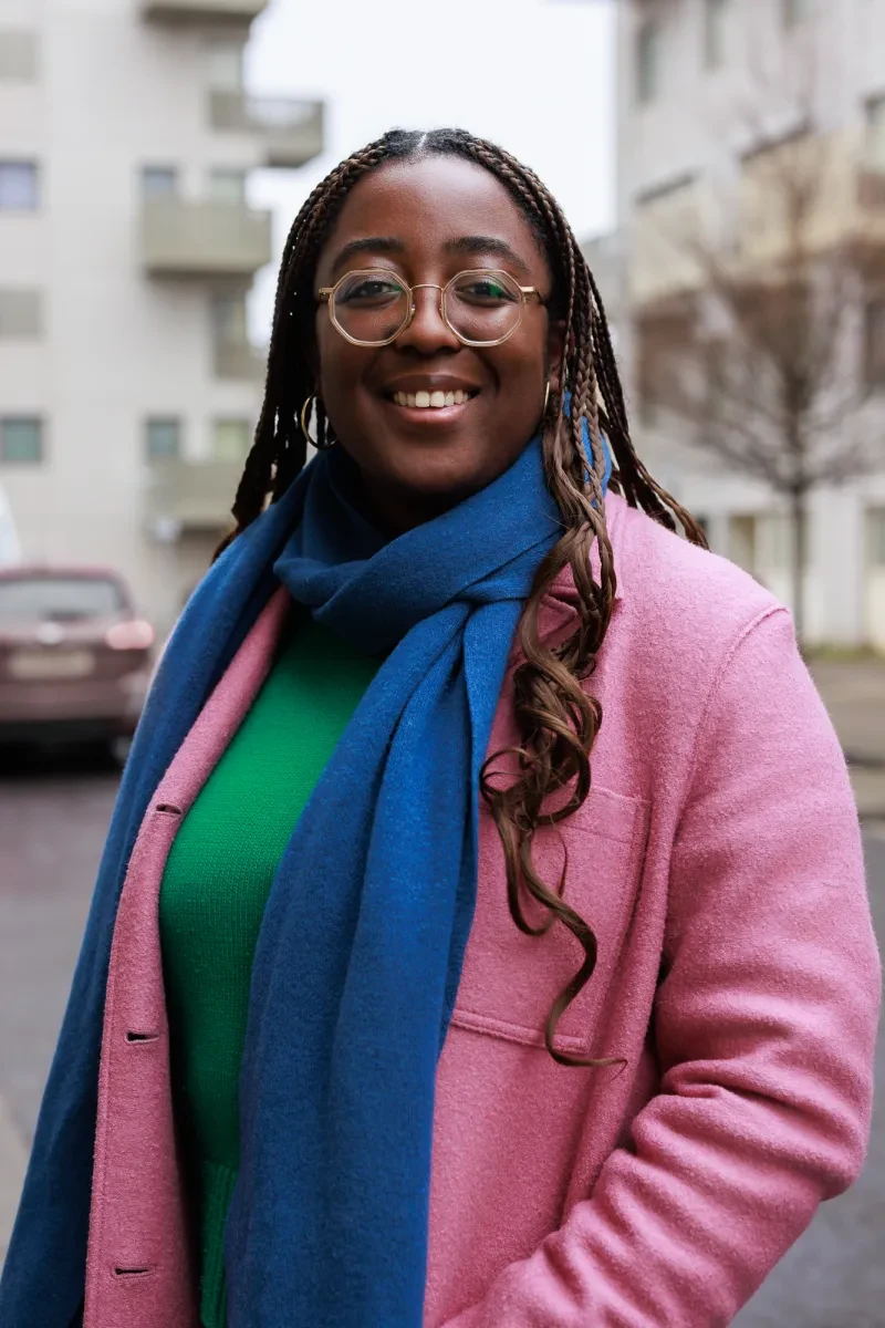 Betty Owoo smiling at the camera, wearing glasses, a blue scarf, green jumper and pink coat