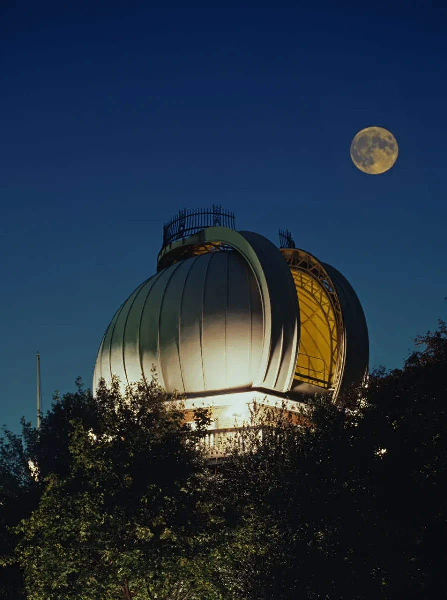 The Great Equatorial Telescope dome of the Royal Observatory Greenwich, lit up at night with a full moon in the sky above