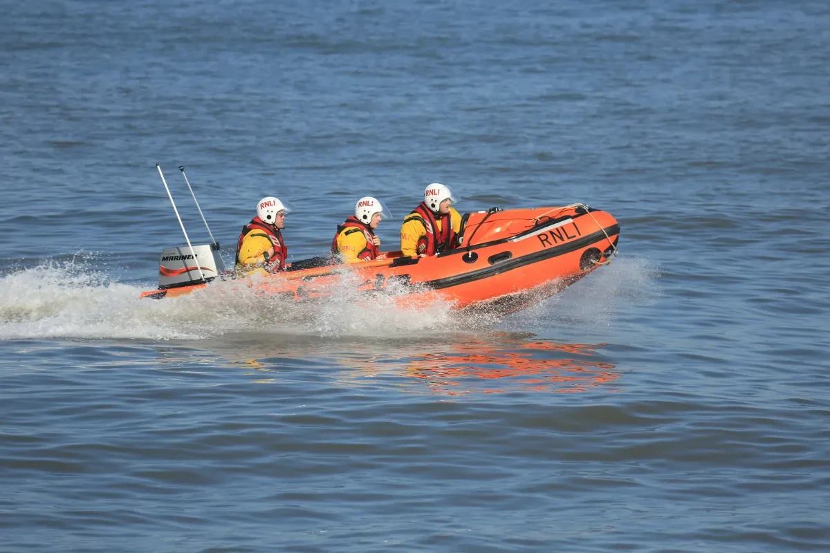 Image showing a bright orange RNLI boat moving fast on a flat blue sea, with three crew members in full yellow heavy gear and white helmets on