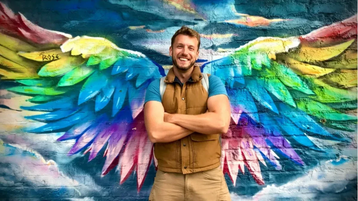 Sacha Coward stands with his hands crossed, wearing a brown gilet and blue shirt. He stands in the centre of some rainbow coloured wings painted on the wall