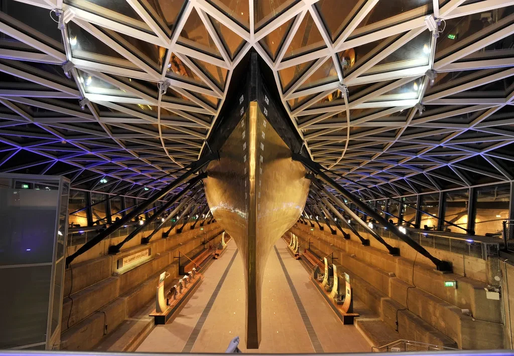 Photo of a large copper hull of a ship in a 'Dry Dock', a dock with stepped sides and supports holding up the ship. The roof is made up of loads of triangular glass panels.
