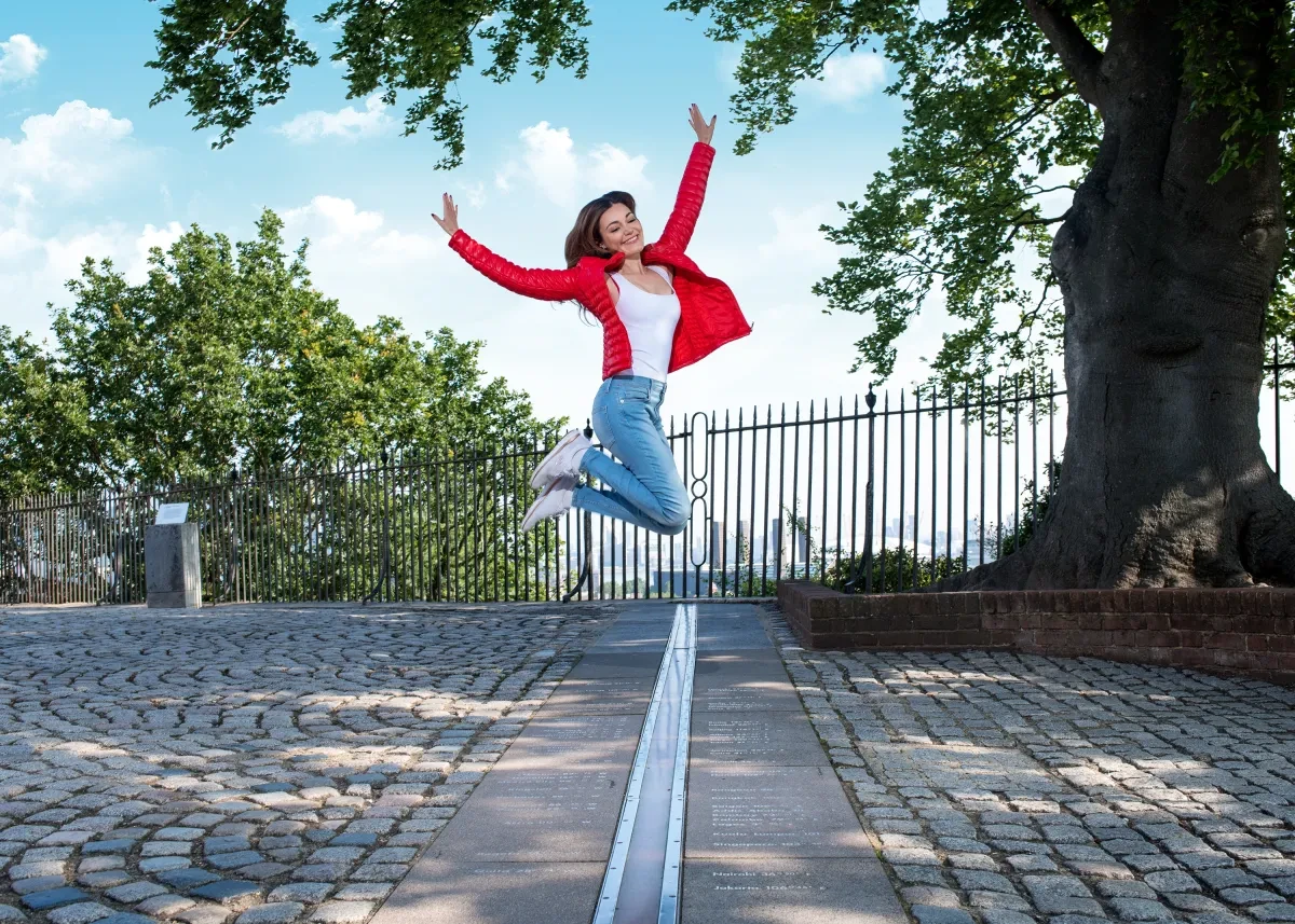 A woman in a red jacket leaps across the Prime Meridian Line at Royal Observatory Greenwich
