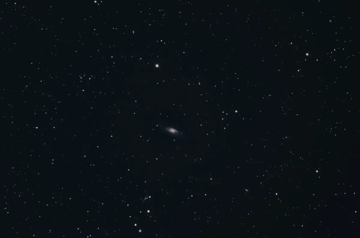 Image showing a dark black sky with multiple stars dotted around, in the centre of the image is a distant white oval shaped smudge which is a galaxy M106, of which it is possible to make out a dark white core with blue and purple spiral arms coming off of it