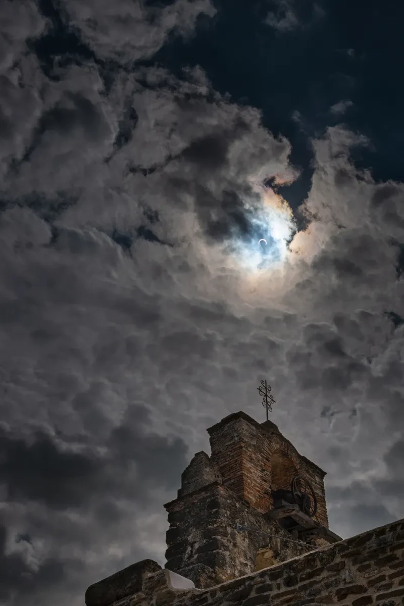 Image from down below looking up at a old stone bell tower with a weathervane on top, in the sky behind there is very dense clouds which thin out towards the top of the image to reveal a dark blue sky. Shining through a small patch of the cloud is the Sun with the moon covering it surface almost fully.