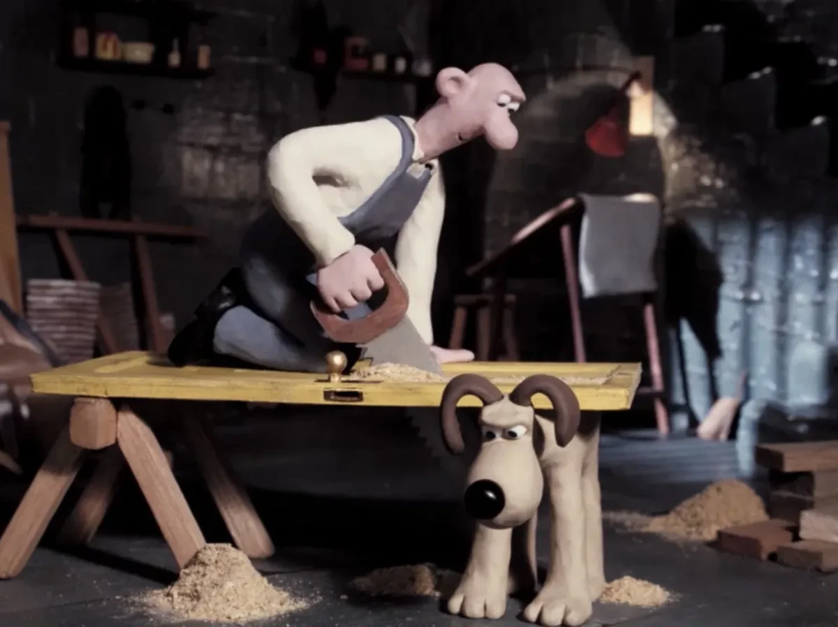 Still from Wallace and Gromit film A Grand Day Out where Wallace, a clay man, is kneeling on a door and sawing it propped up on a bench and also by Gromit, his dog