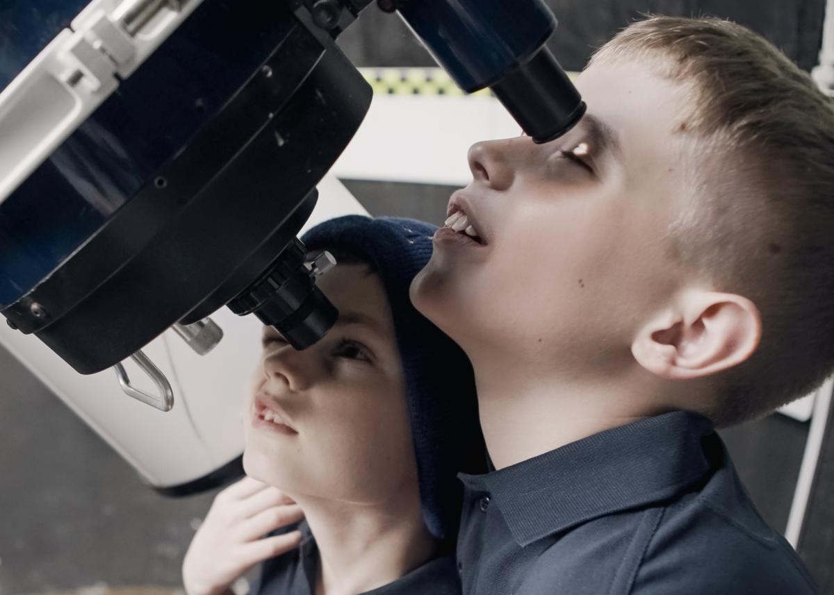 Photo of two young brothers looking up into the lenses of a large telescope, the one on the right has his arm around the other