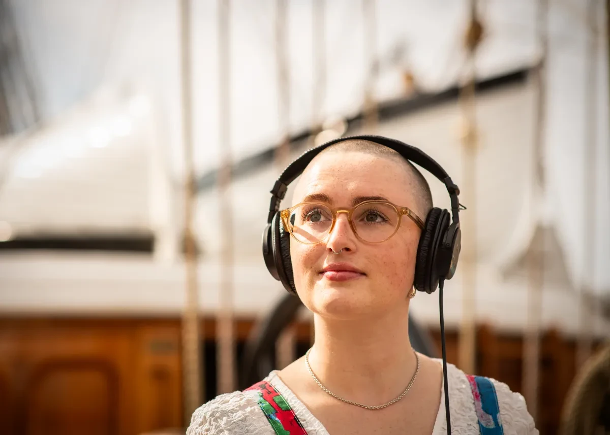 A woman wearing glasses and with a close-shaven haircut looks wistful as she listens to something on headphones