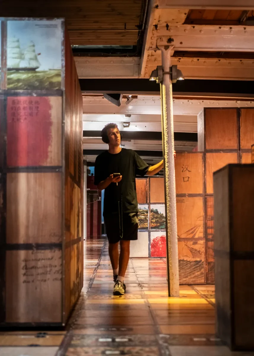 A young man walks among wooden crates in the lower hold of Cutty Sark. He is wearing headphones and holding his phone in his right hand