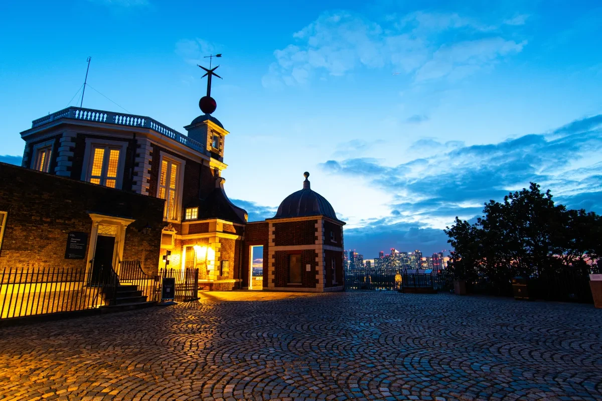 The Royal Observatory's historic Flamsteed House building lit up at dusk, with the lights of Canary Wharf visible in the distance