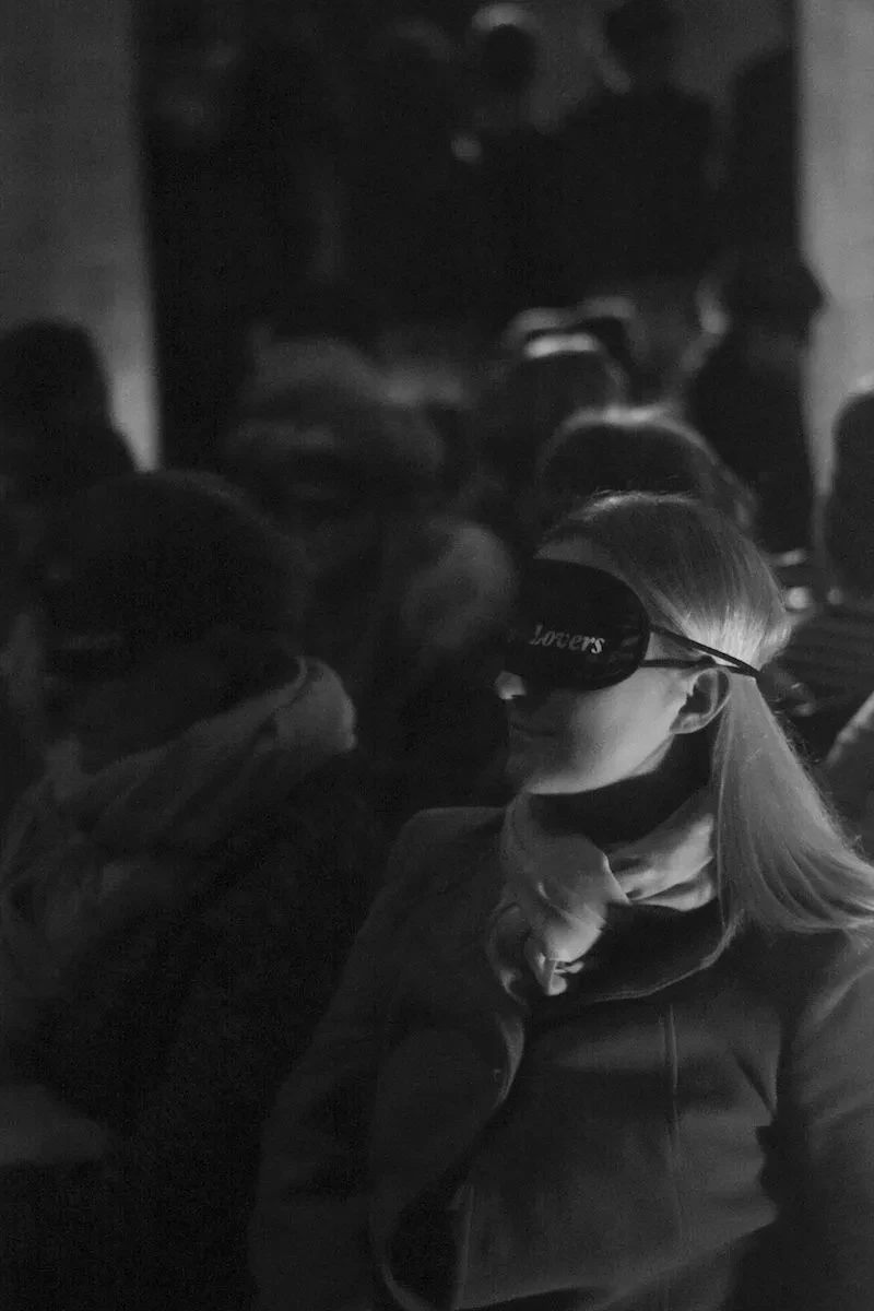 Darkened photo where it's possible to make out people sitting down in rows wearing blindfolds