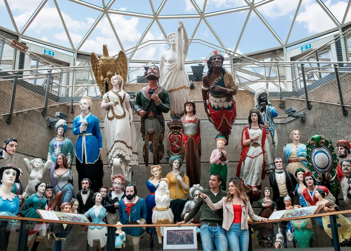 A couple stand in front of a display of ship figureheads. A whole range of characters and designs are arranged, positioned as they would have appeared on the front of ships