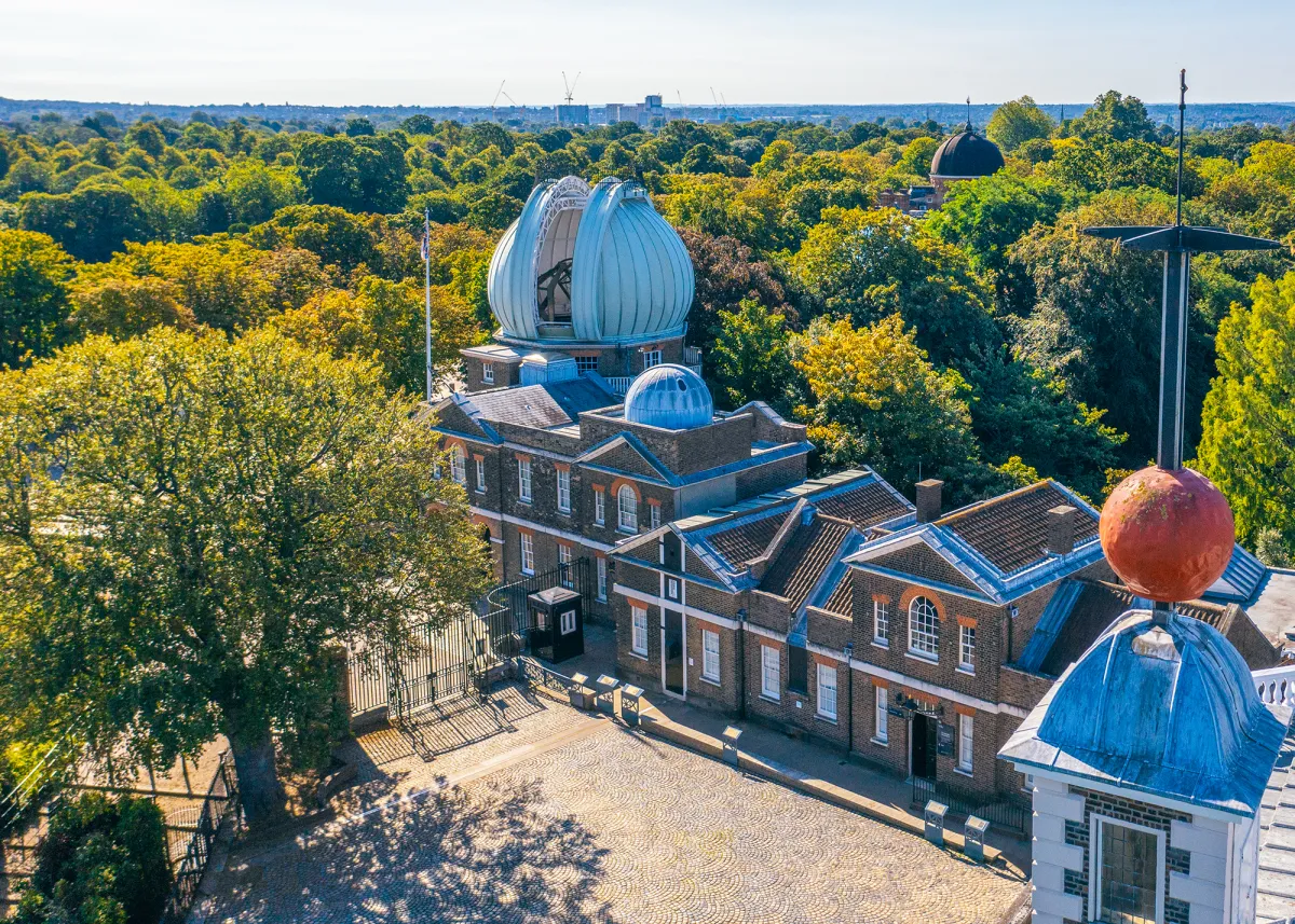 Drone view of the historic Royal Observatory buildings during the day. Trees surround a central group of brick buildings, with the distinctive 'Onion Dome' of the Great Equatorial Telescope in the centre