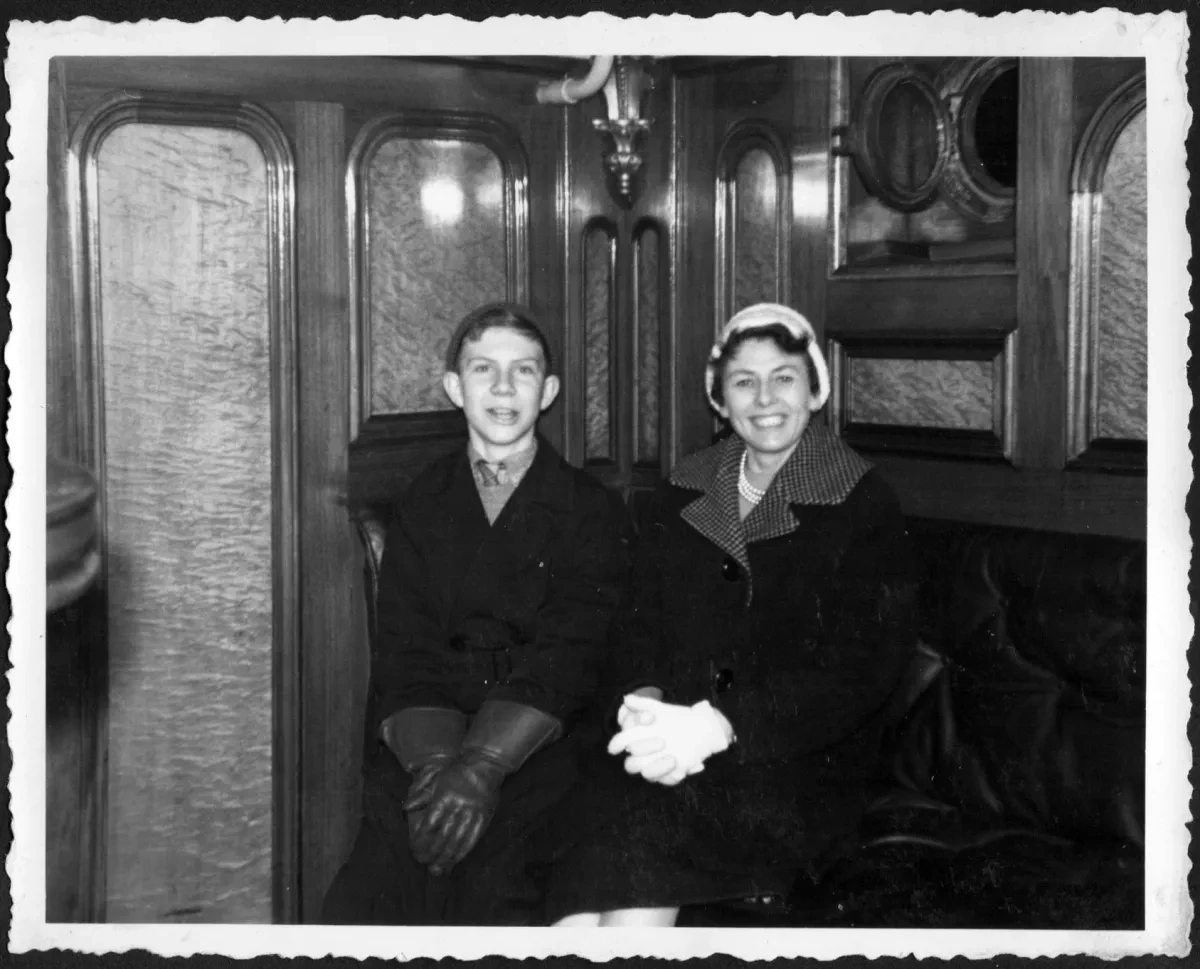 Black and white photograph of a boy with his mother, smiling brightly in in a wood-panelled saloon.