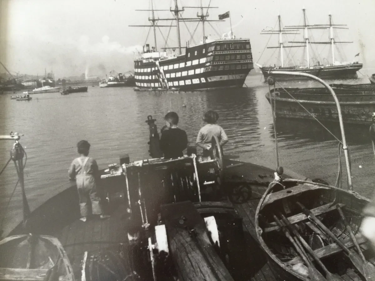 Black and white photograph showing three children standing at the stern of a boat, looking out at ships in the River Thames.