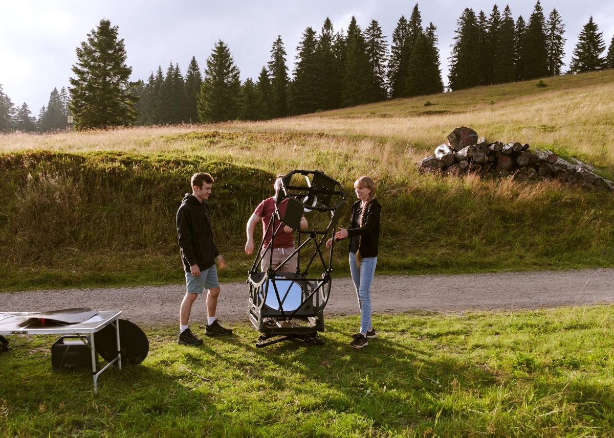 Landscape photo of three people on a golden green Austrian mountain standing around a large telescope which has a big metal frame and is around 5.5 feet tall