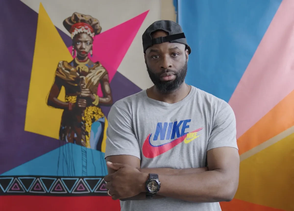 Artist Joseph Ijoyemi stands in front of an artwork of a queen on a backdrop of colourful triangles